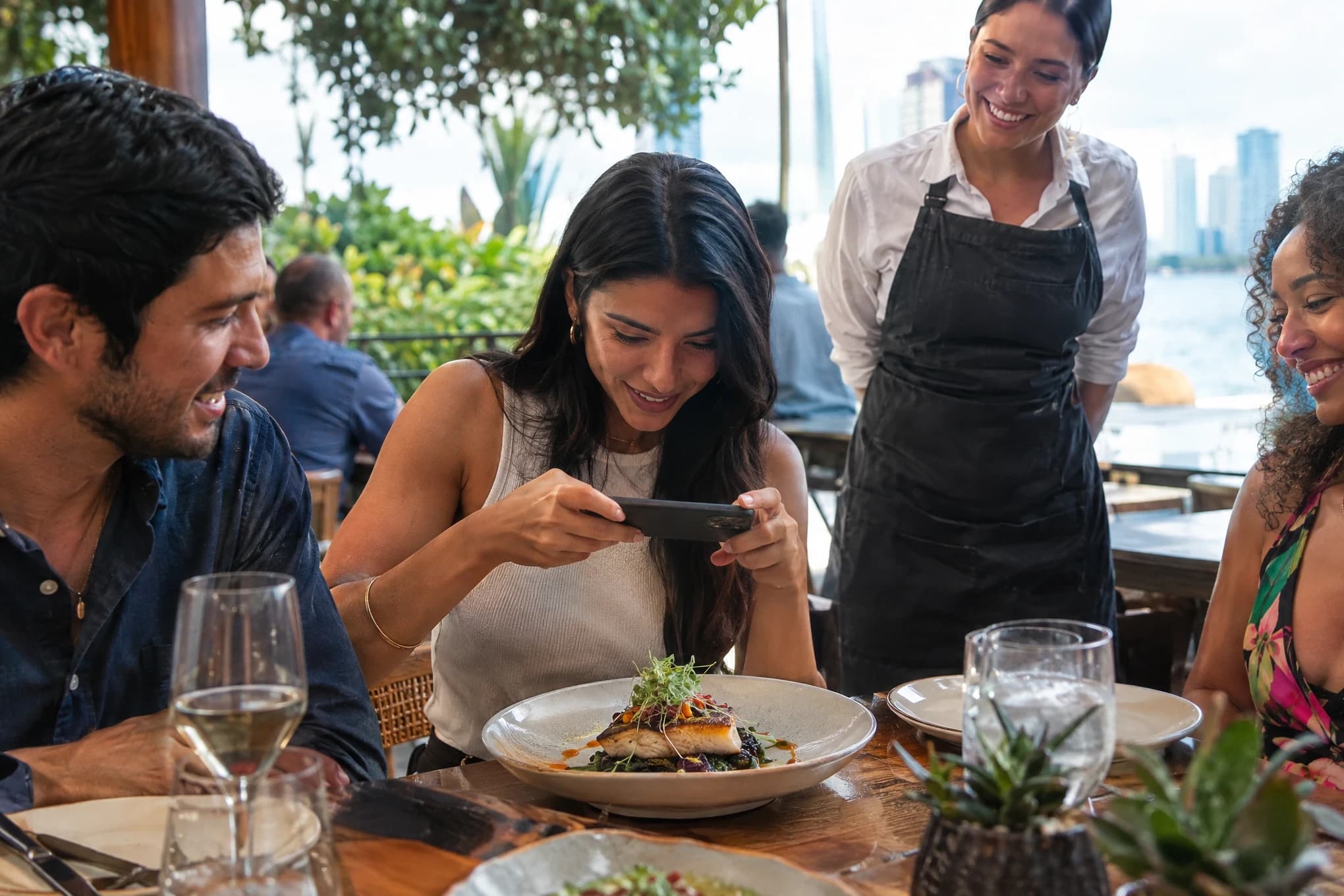 Friends at a restaurant table; one photographs a dish with her phone while the server smiles