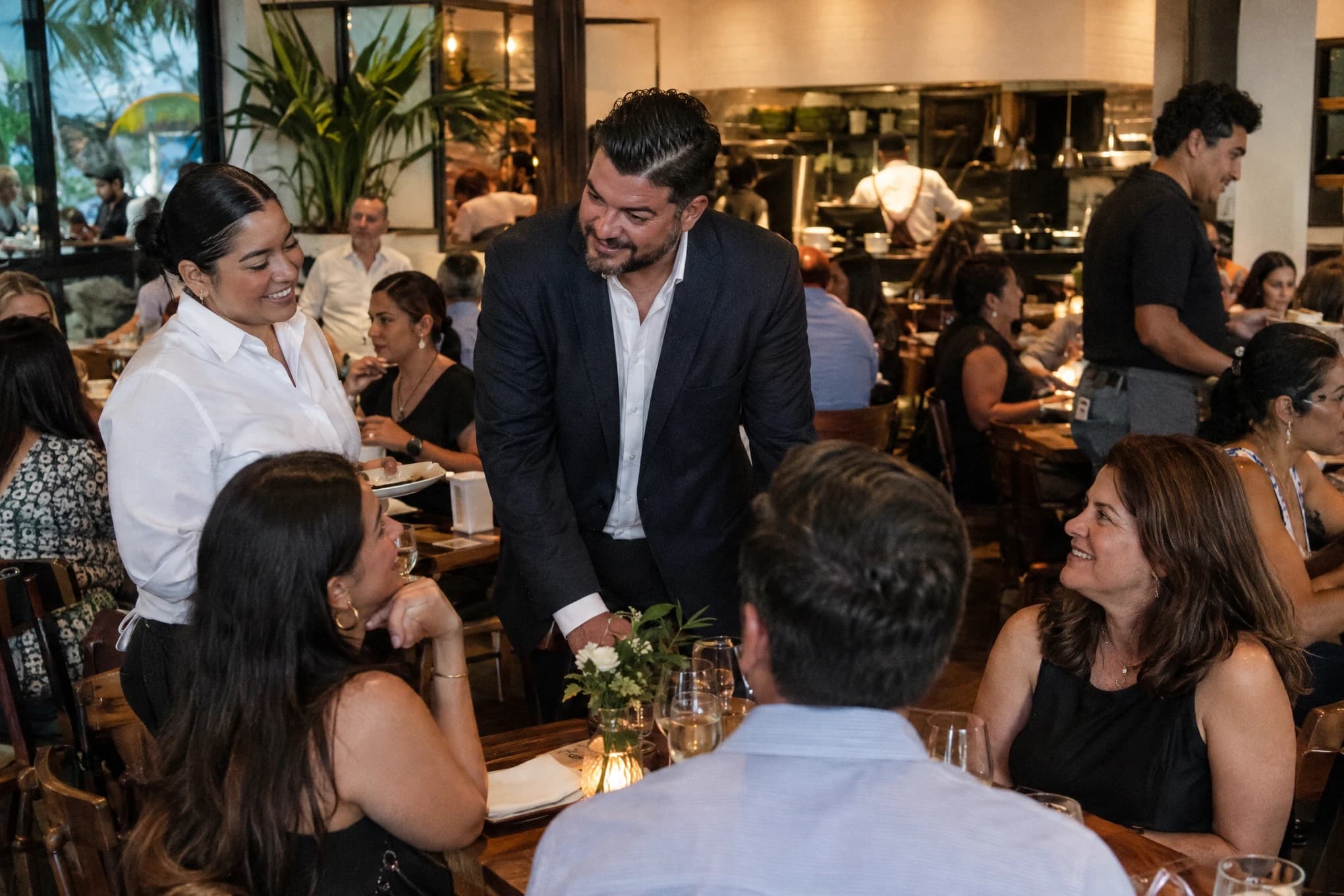 Clientes conversan con meseros y el gerente de un restaurante concurrido durante la cena
