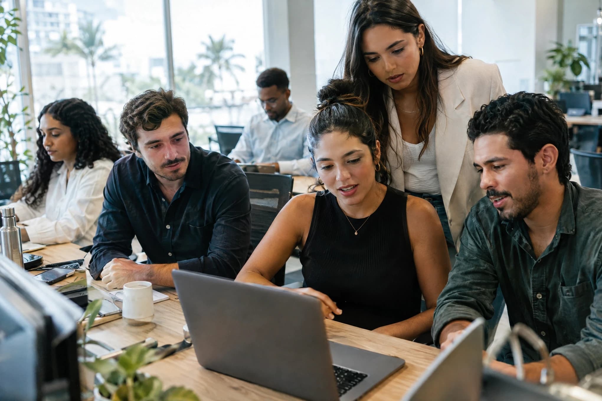 A six-person agency team works at shared desks while a client looks at a blurred laptop screen