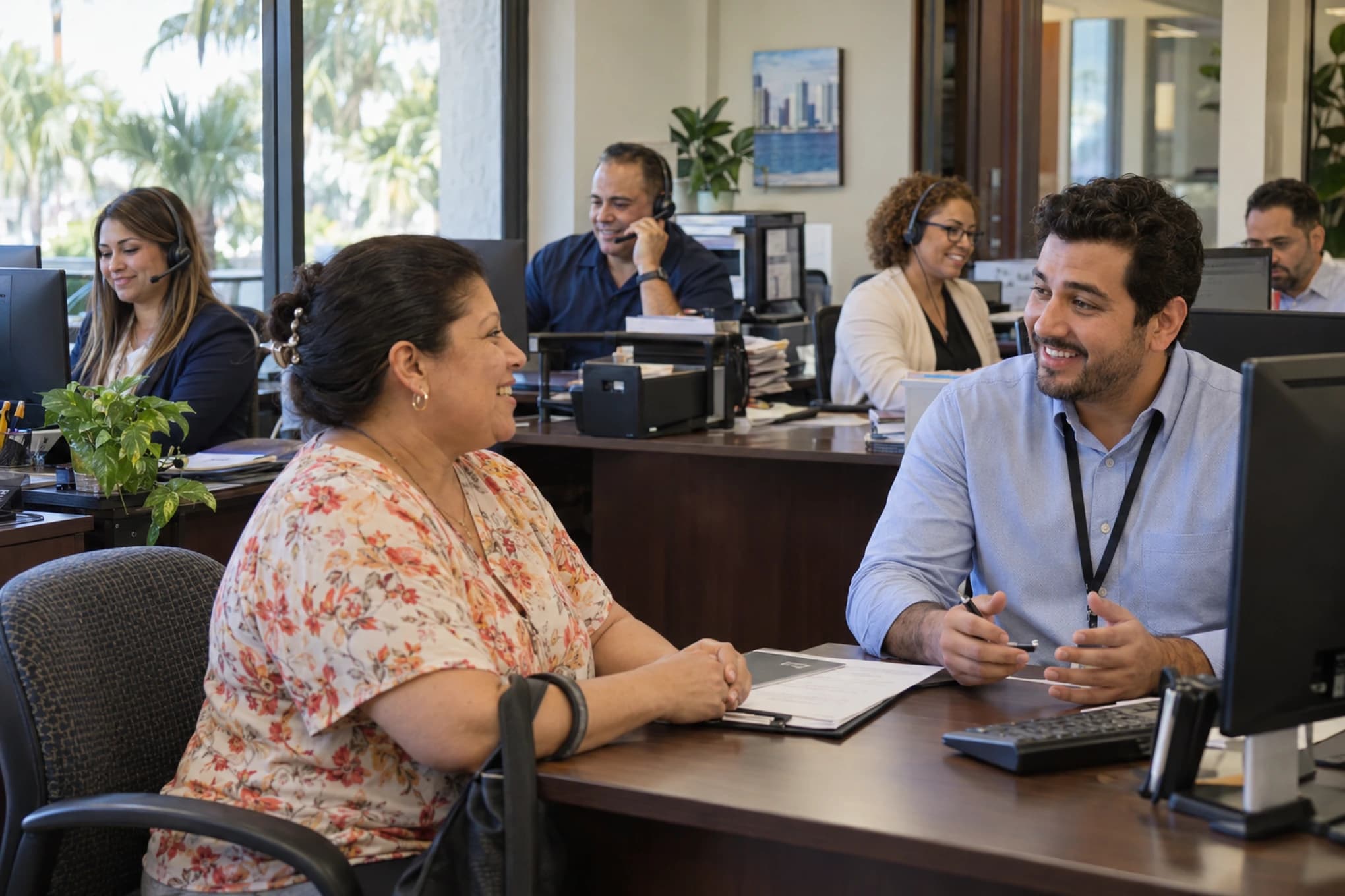 Cinco empleados bilingües en el área de atención de una oficina de servicios trabajan frente a sus monitores con auriculares, uno de ellos conversa con una clienta sentada frente a su escritorio