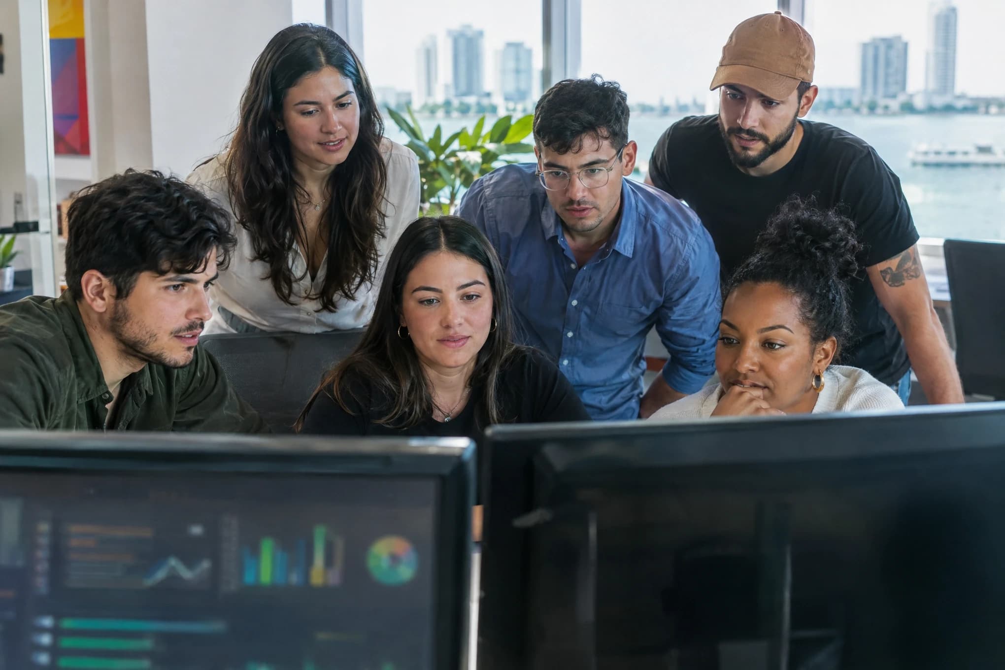 A six-person ad team launches a campaign in front of two monitors with blurred dashboards