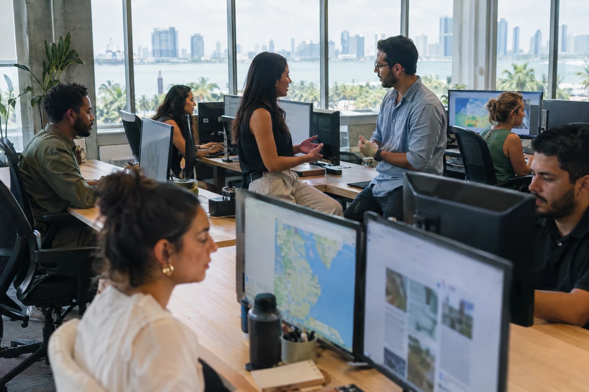 A six-person SEO team works at long desks with blurred maps and pages on their monitors