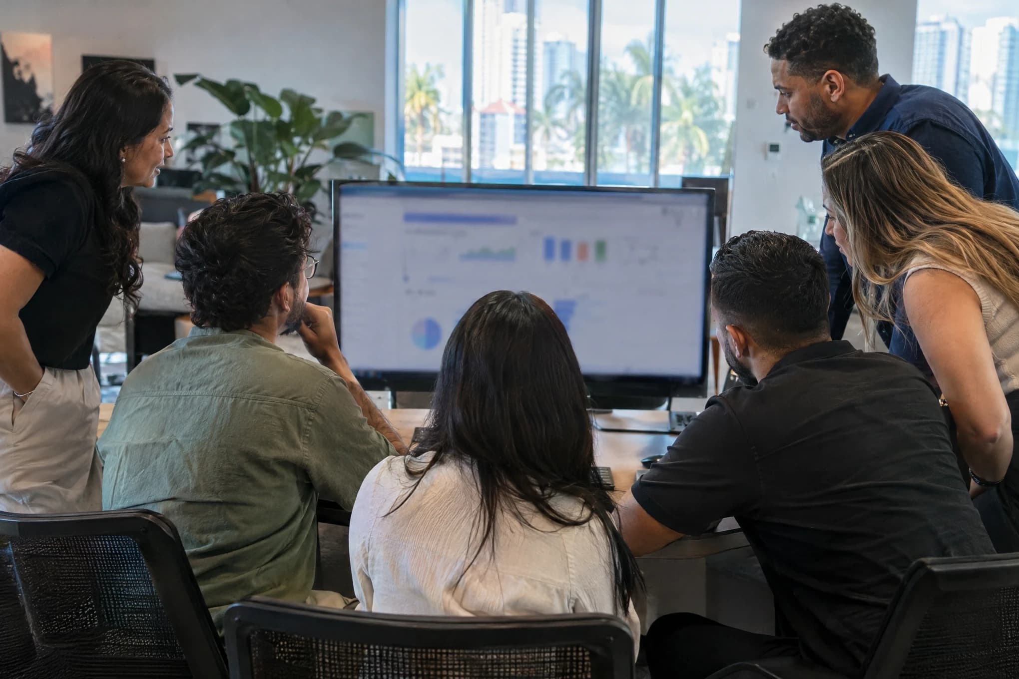 Cuatro especialistas en anuncios y dos ejecutivos de cuenta revisan un tablero desenfocado en un monitor grande