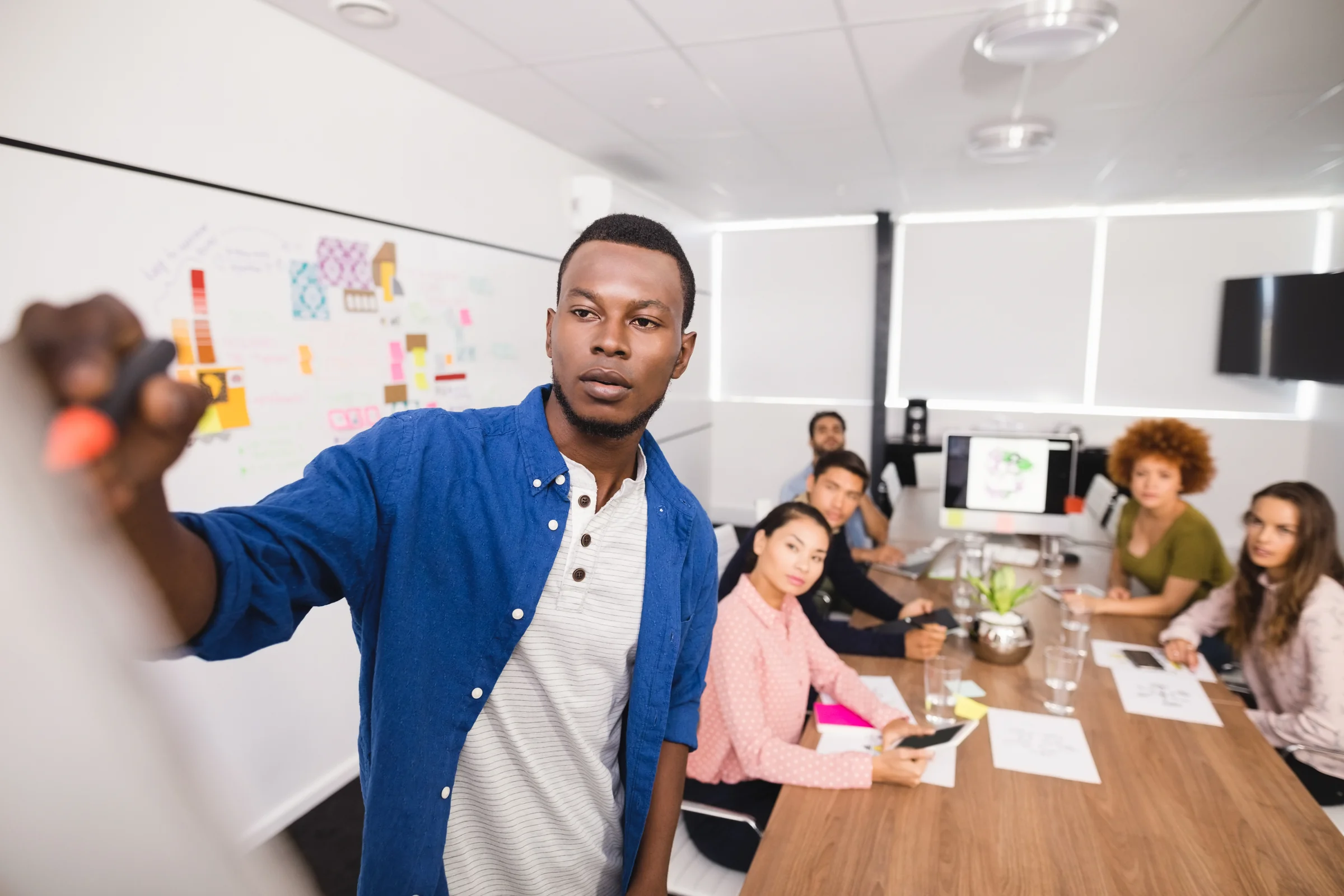 A manager explains a process on a whiteboard with colored notes to a team seated around a meeting table