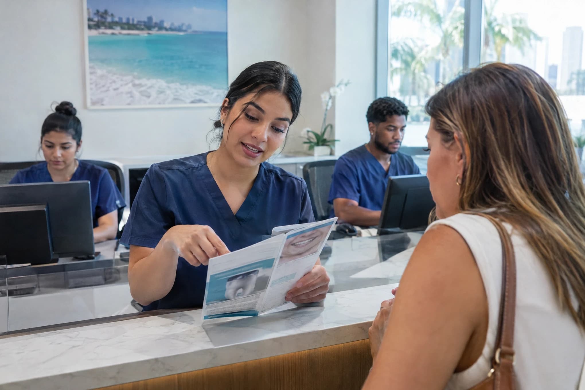 At a dental clinic front desk, a receptionist explains a brochure to a patient while two colleagues work behind her