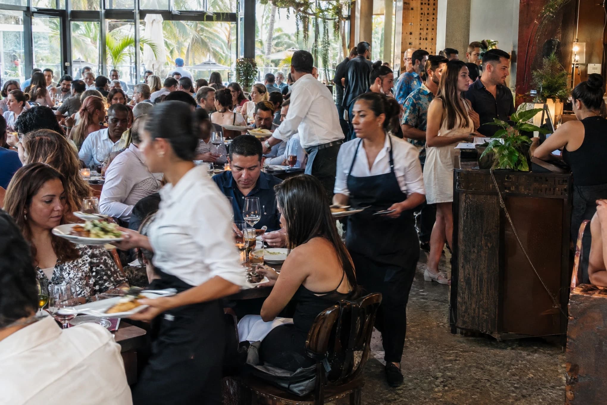 A packed restaurant at peak hour, servers rushing and a line of guests waiting