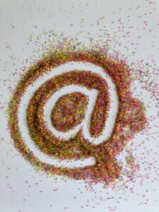 Imagen artística del símbolo arroba (@), creado con brillantes cuentas o partículas multicolores, sobre fondo blanco. Representa conceptos clave como conexión digital, identidad online, email marketing, redes sociales y presencia en internet.