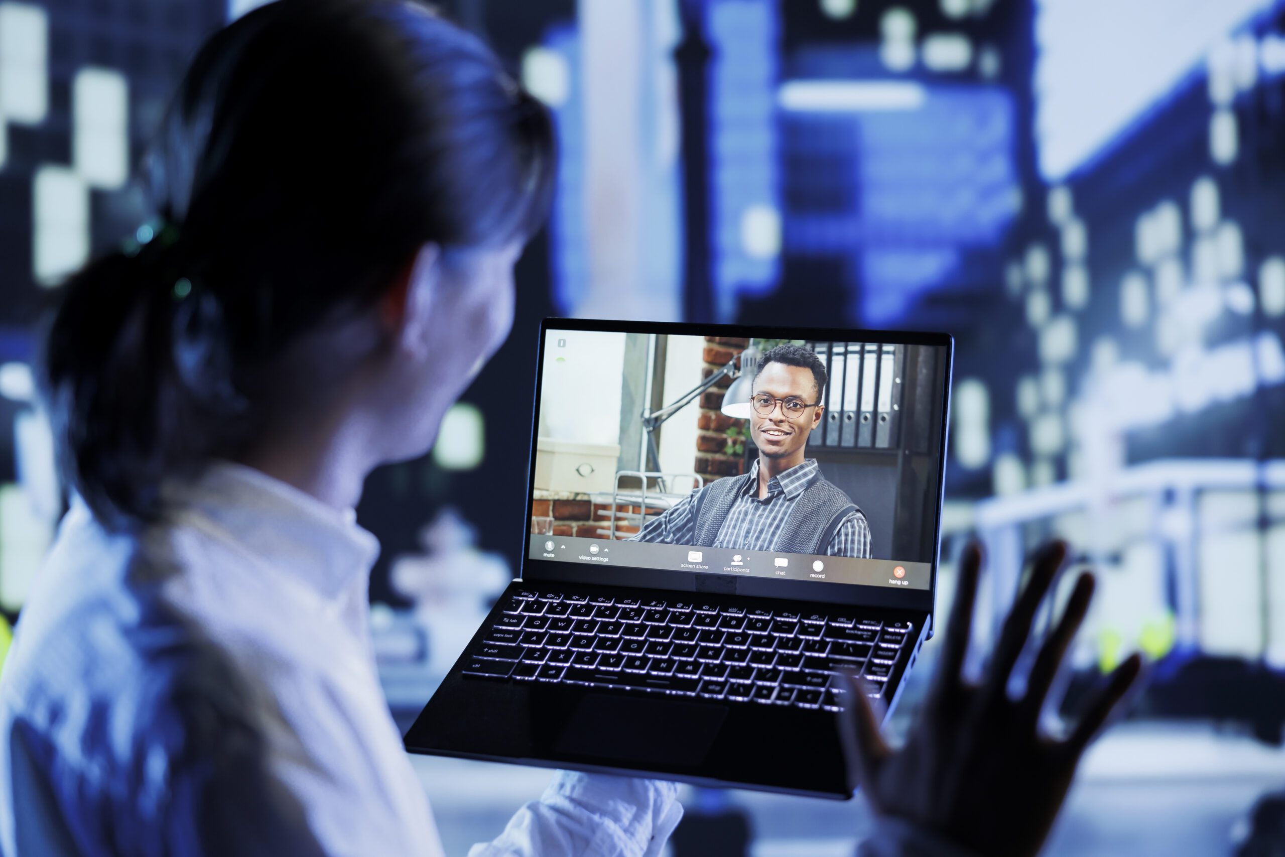 Asian man in videocall with coworker while strolling around city streets at night, giving him work updates. Person using laptop to show colleague surroundings in nighttime urban environment